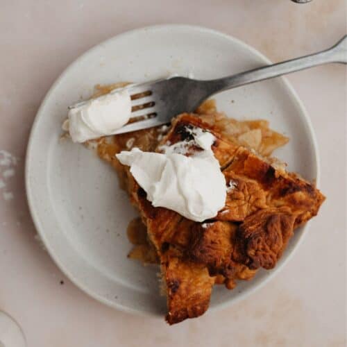 A slice of bourbon apple pie on a small plate. The pie has a dollop of whipped cream