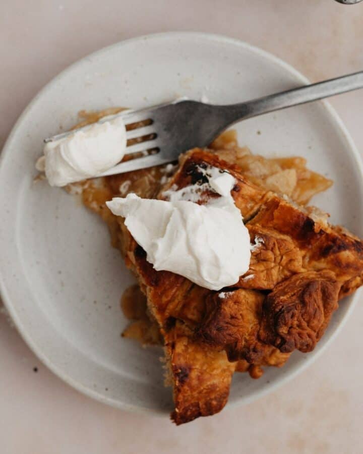 A slice of bourbon apple pie on a small plate. The pie has a dollop of whipped cream