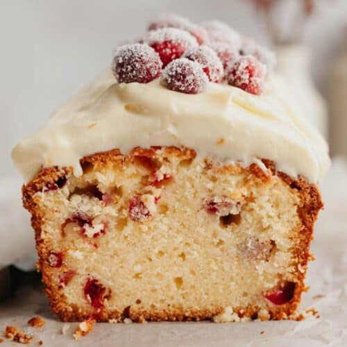 A cranberry loaf cake cut in half showing the inside. It is topped with white frosting and sugared cranberries.