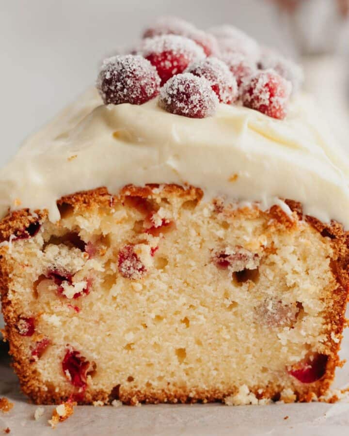A cranberry loaf cake cut in half showing the inside. It is topped with white frosting and sugared cranberries.