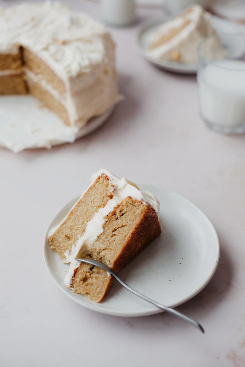 A slice of cardamom cake with a white frosting on a small plate with a spoon. You can see the rest of the cake on parchment paper behind, with a glass of milk