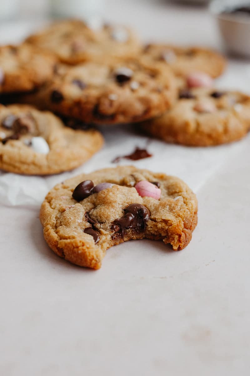 A chocolate chip cookie with a bite taken out of it. A pile of cookies are behind it on parchment paper
