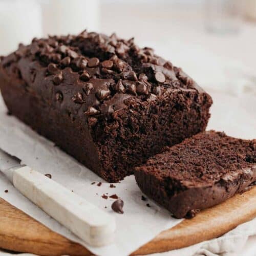 A chocolate loaf cake on parchment paper topped with chocolate chips, one slice has been cut.
