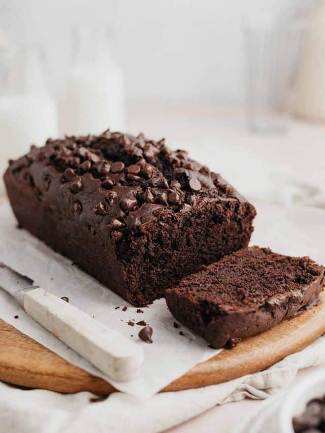 A chocolate loaf cake on parchment paper topped with chocolate chips, one slice has been cut.