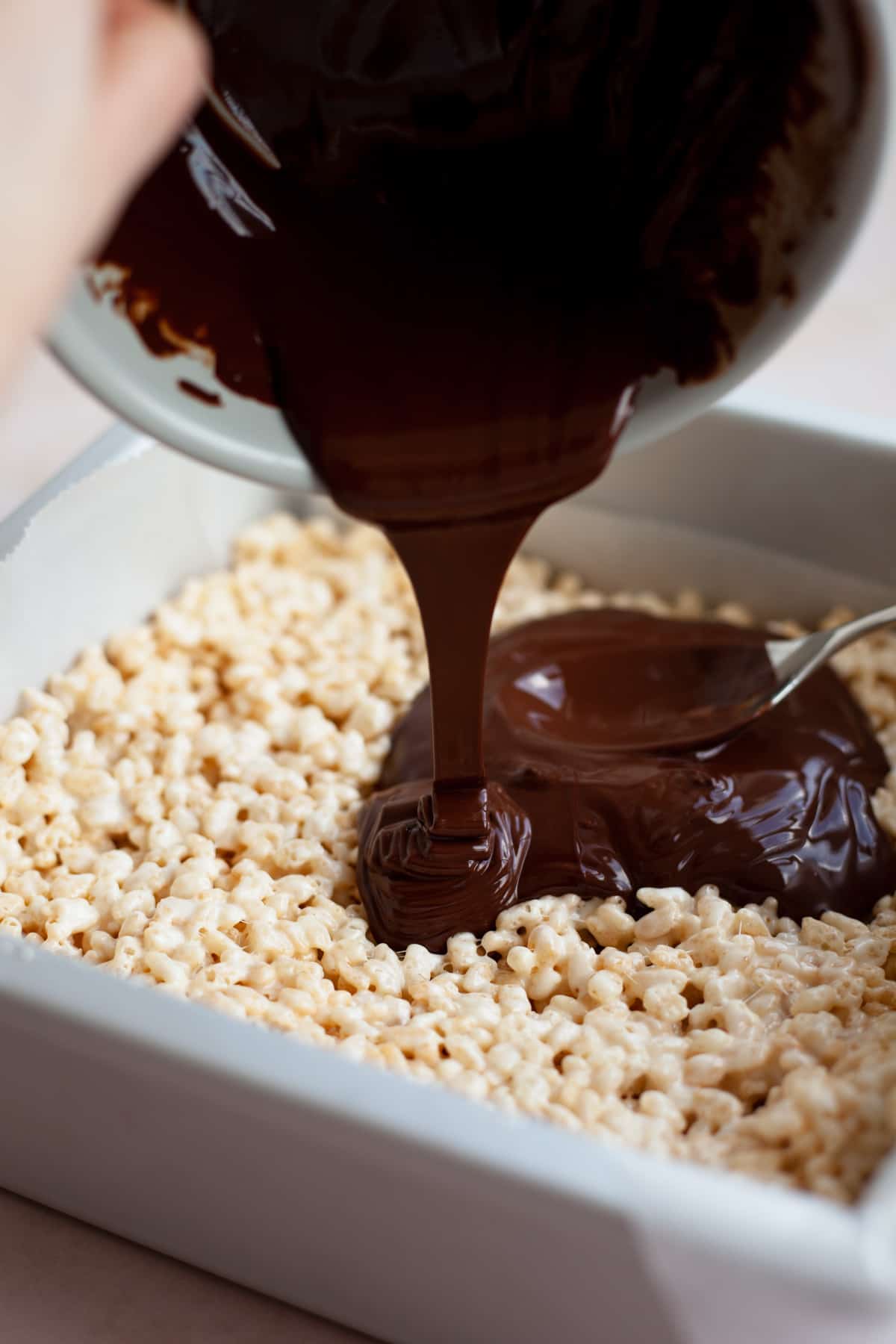 Rice krispie treats packed down into an 8x8 pan, melted chocolate is being poured on top.