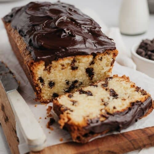 A chocolate chip loaf cake covered in chocolate ganache on a wooden board, one slice has been cut.