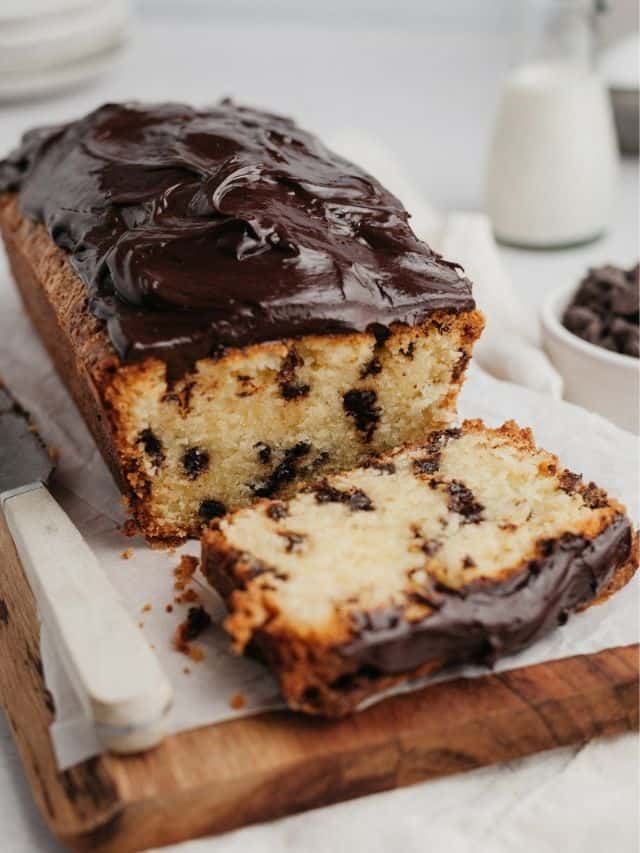 A chocolate chip loaf cake covered in chocolate ganache on a wooden board, one slice has been cut.