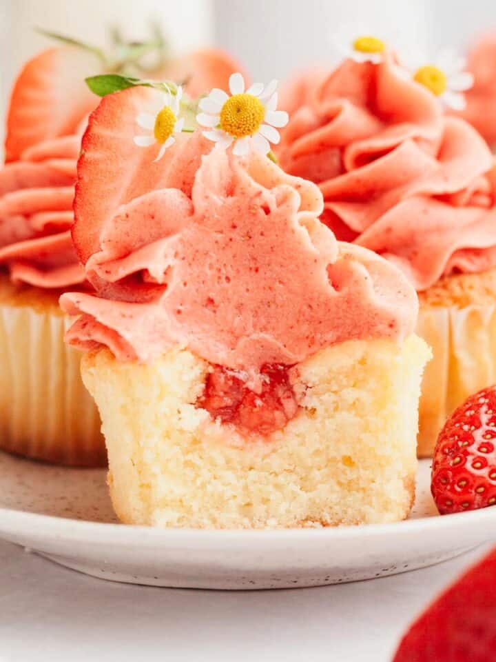 A vanilla cupcake with strawberry filling and strawberry frosting, a bite has been taken out of it.