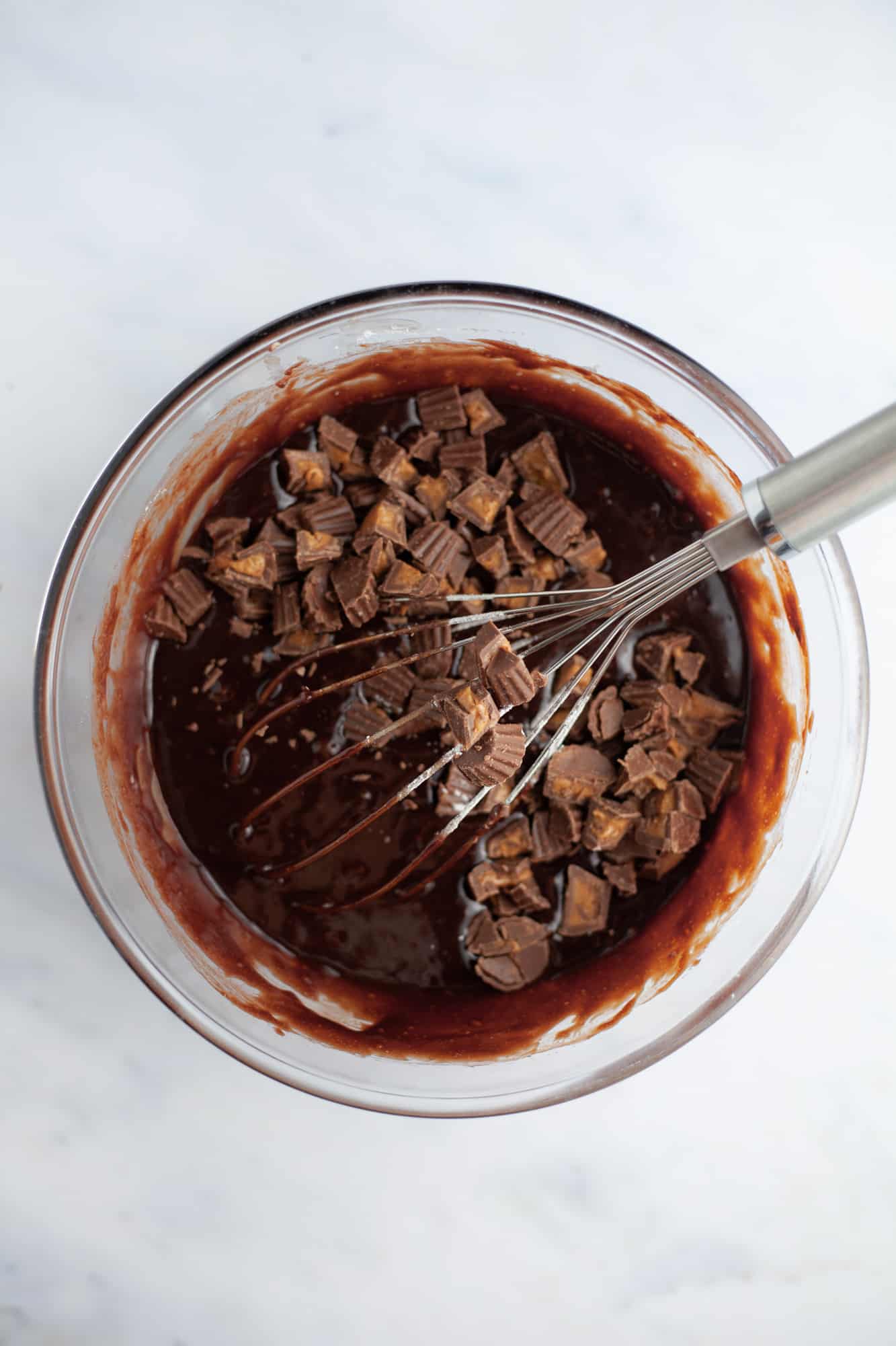 A pyrex bowl with brownie batter and chopped peanut butter cups and a large silver whisk