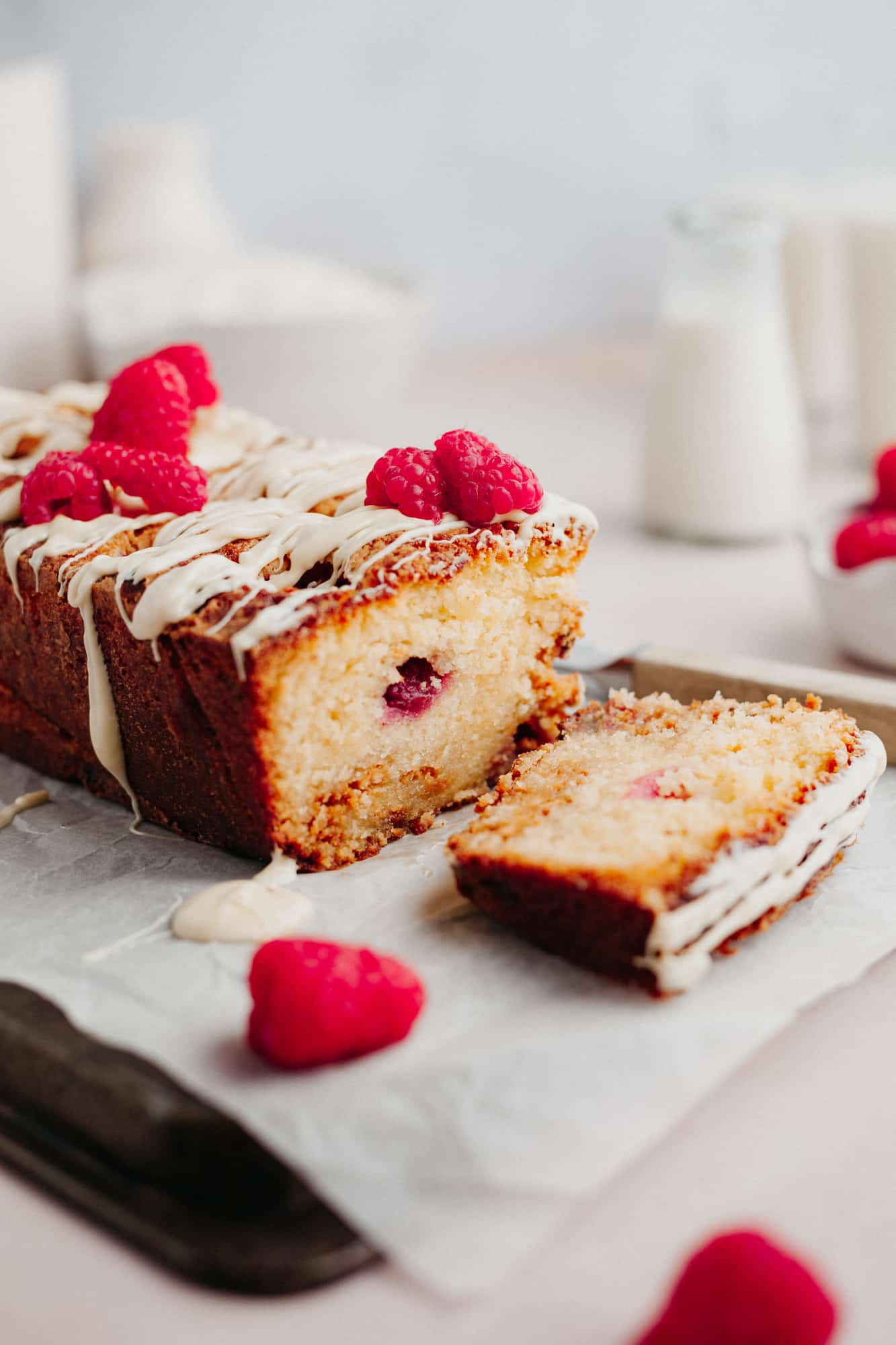 A raspberry and white chocolate loaf cake topped with white chocolate drizzle and fresh raspberries. One slice is cut.