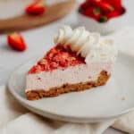 A slice of strawberry cheesecake with a whipped cream topping on a small beige plate. You can see strawberries in a ceramic bowl in the background