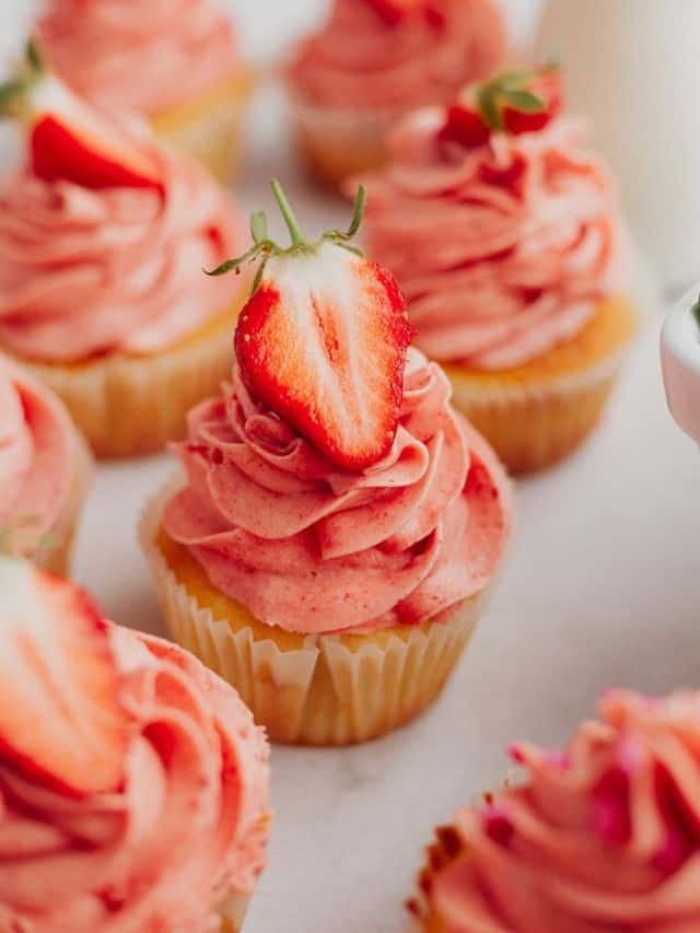 A close up of a vanilla cupcake frosted with pink frosting, with half a strawberry placed on top