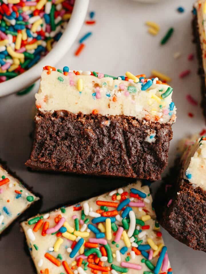 Brownies with an edible cookie dough layer with rainbow sprinkles on parchment paper.