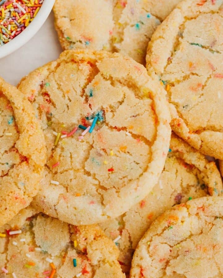A pile of sprinkle sugar cookies with a small bowl of sprinkles.