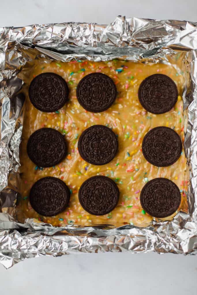 A brownie pan lined with foil and an unbaked layer of funfetti cake, and rows of oreos
