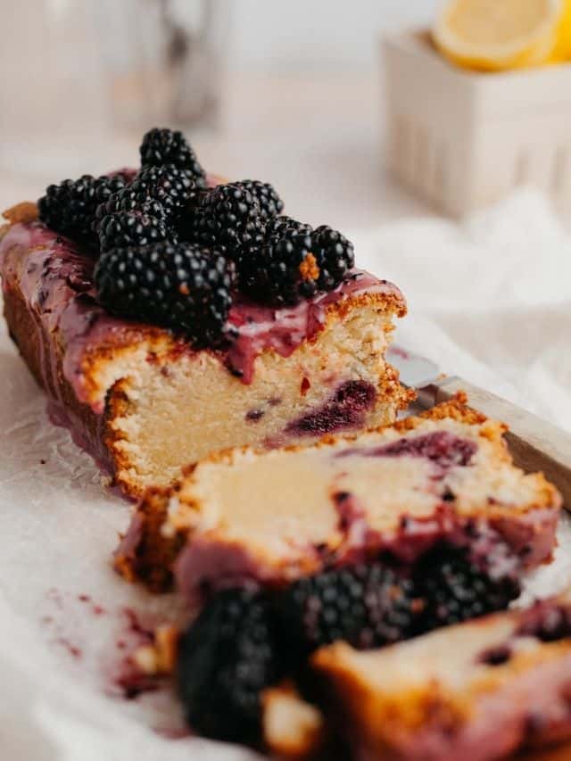 A lemon blackberry loaf covered in a purple glaze and blackberries. Several slices have been cut off.