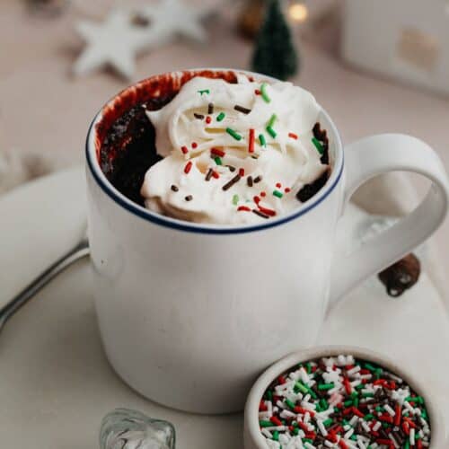 A red velvet mug cake topped with whipped cream and red and green sprinkles.
