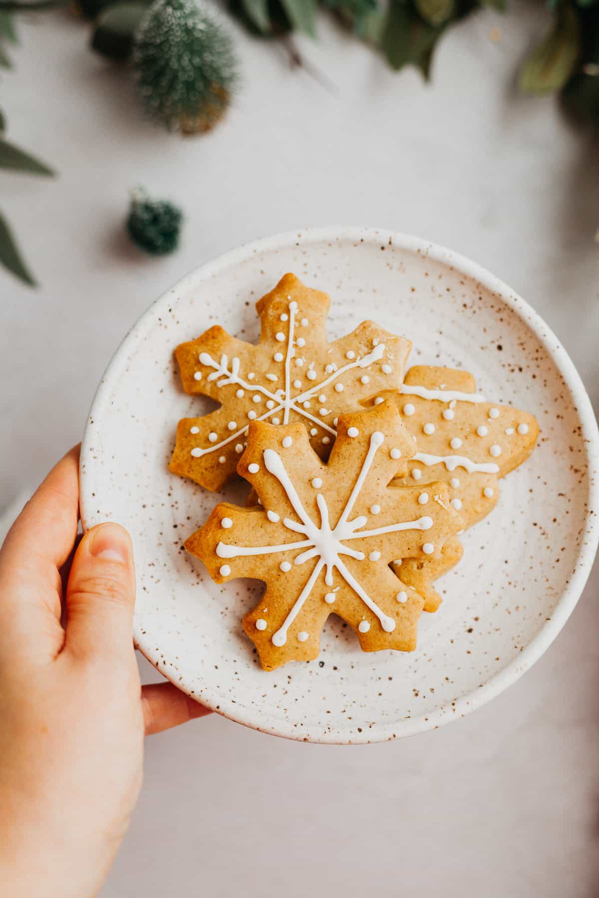 A woman's hand holding a small white plate with three decorated gingerbread cookies in the shape of snowflakes.