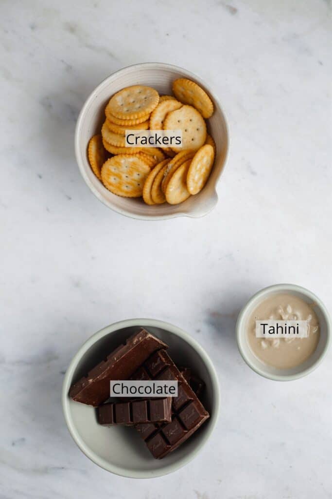 dark chocolate chopped in a bowl, ritz crackers in a bowl, and a small with tahini on a marble counter.
