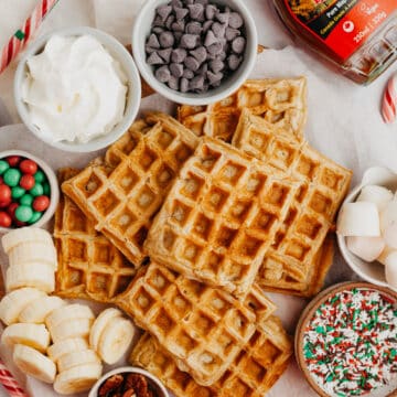 A pile of buttermilk waffles surrounded by sprinkles, chocolate chips, maple syrup and sliced bananas.