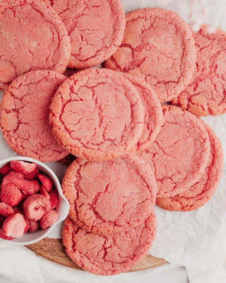 several pink sugar cookies on parchment paper with a small bowl of freeze dried strawberries.