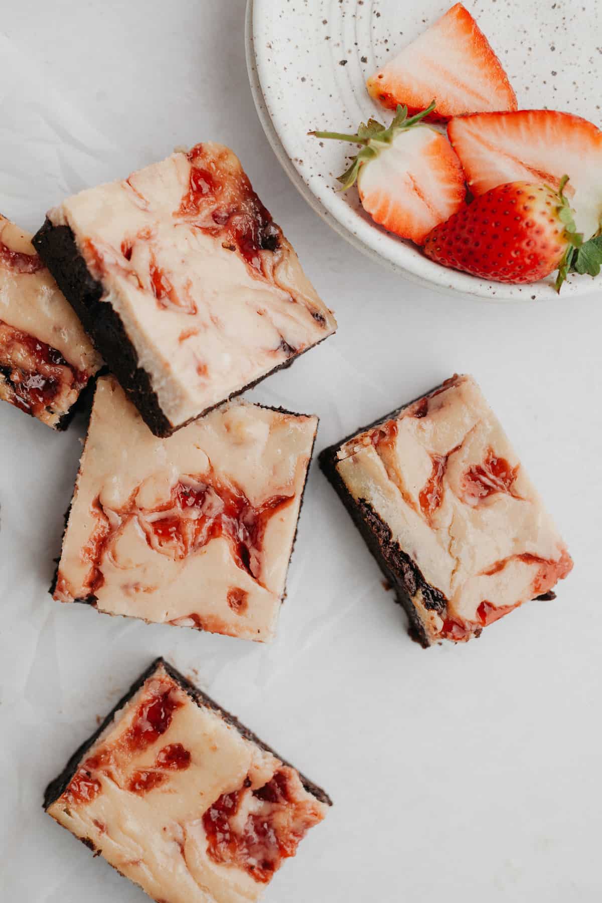 Several strawberry cheesecake brownies on parchment paper with several sliced strawberries on a grey plate.