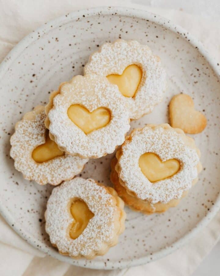 a small speckled plate with lemon curd linzer cookies, there is a heart cutout in each cookie.