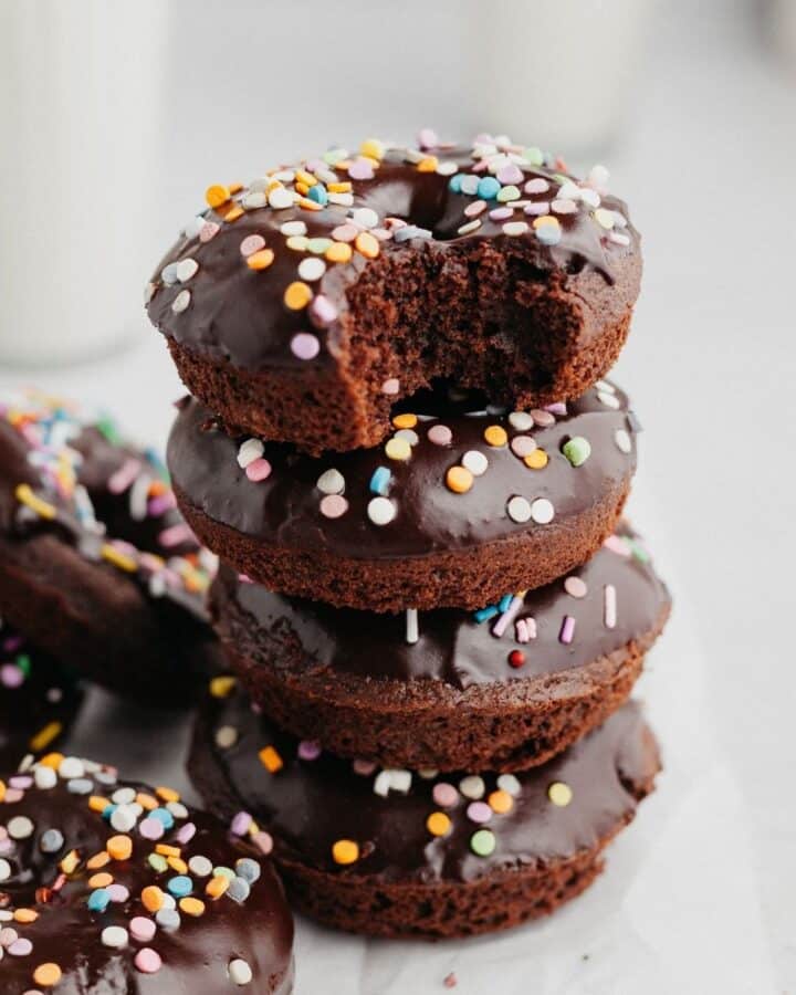 a stack of chocolate sprinkle donuts, the donut on top has a bite taken out of it.