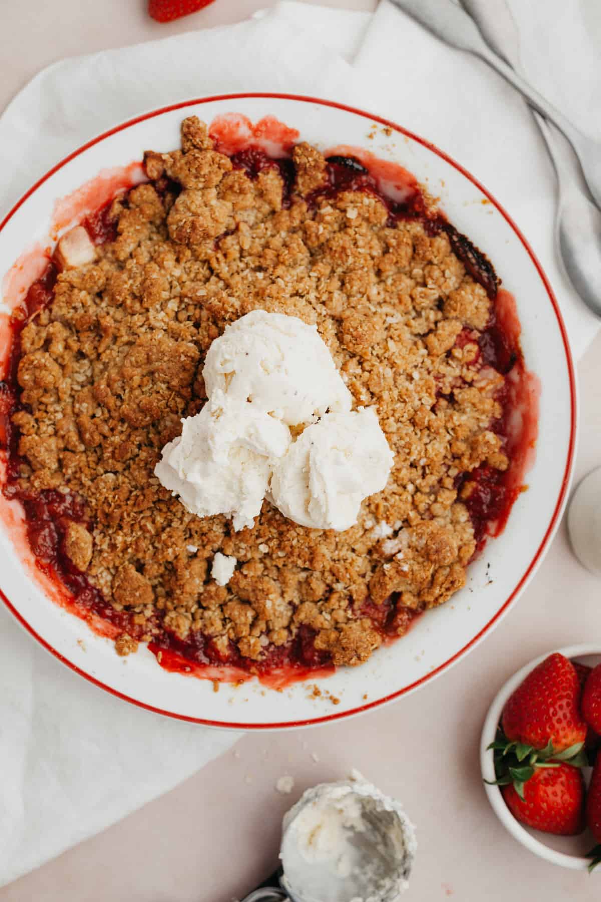 Strawberry and apple crisp in a white pie dish with several scoops of vanilla ice cream in the middle.