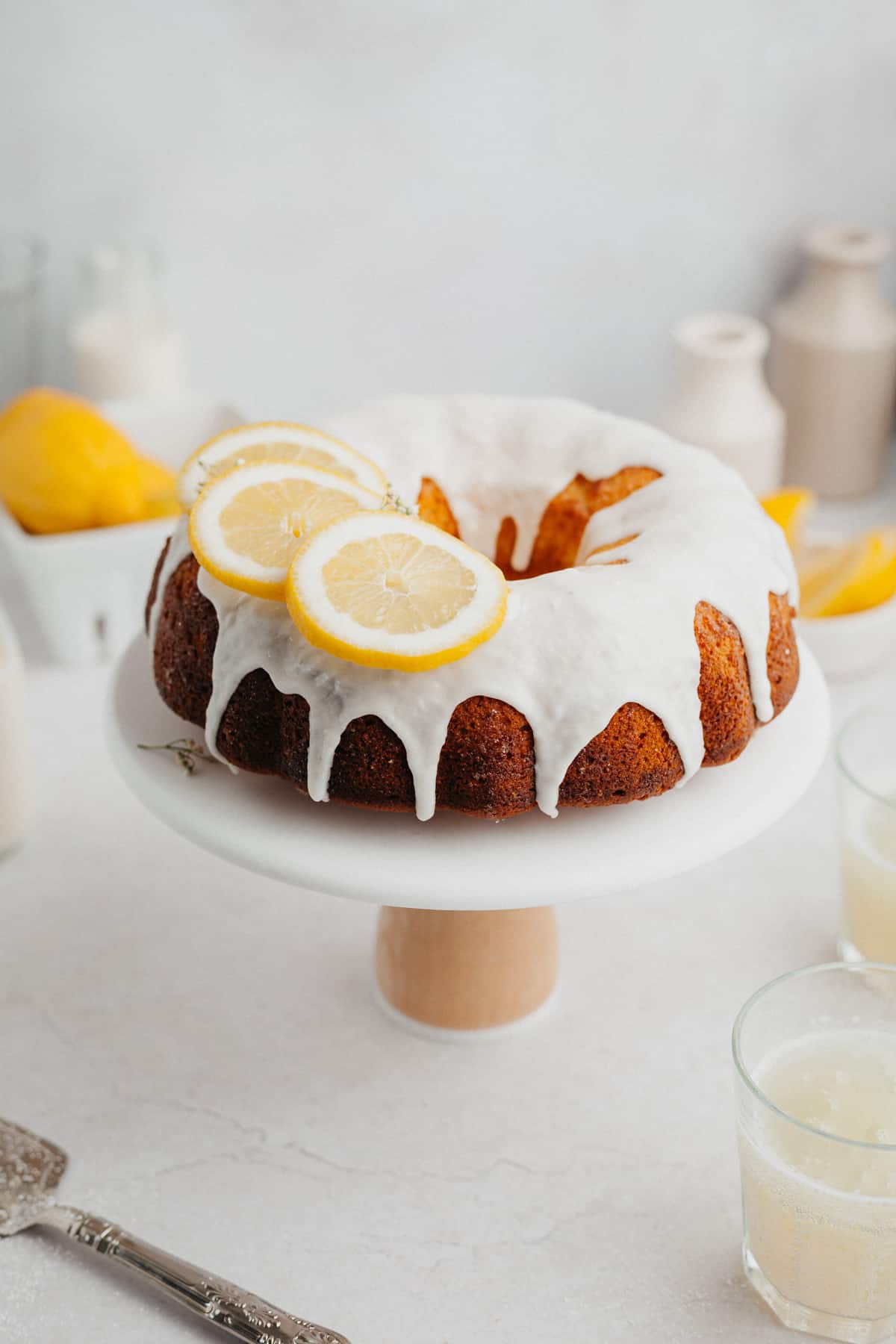 A lemon bundt cake covered in a white glaze and slices of lemon on a white cake stand.