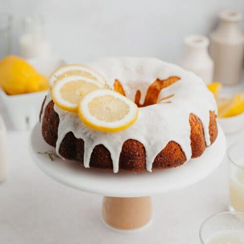 A lemon bundt cake covered in a white glaze and slices of lemon on a white cake stand.
