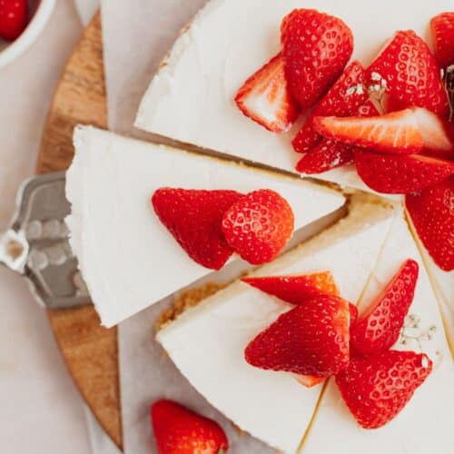 An overhead shot of a white cheesecake topped with sliced strawberries, one slice is being taken out with a silver cake server.