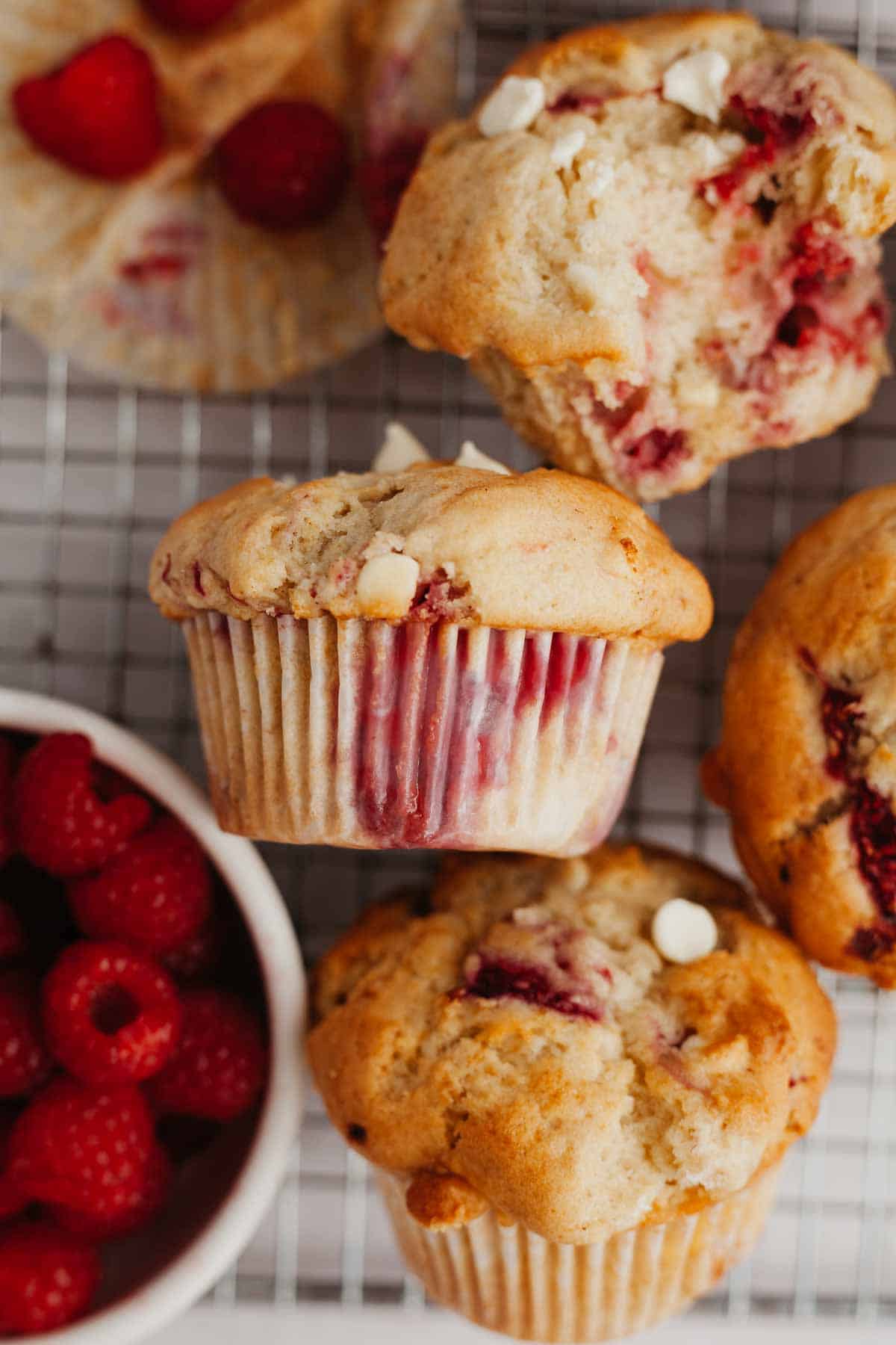 three white chocolate raspberry muffins on a cool rack, one has a bite taken out of it.