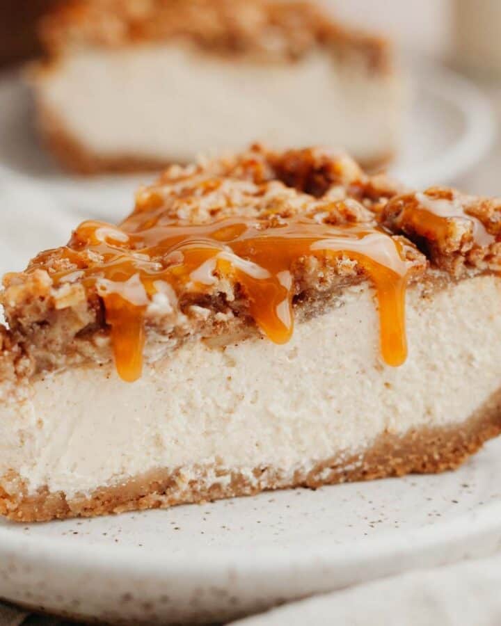 A slice of apple crisp cheesecake drizzled in caramel sauce on a small beige plate.