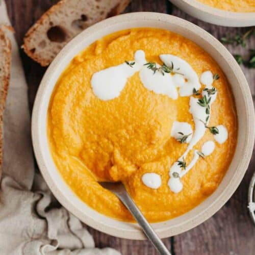 A white bowl with pumpkin carrot soup in it, drizzled with heavy cream and thyme.