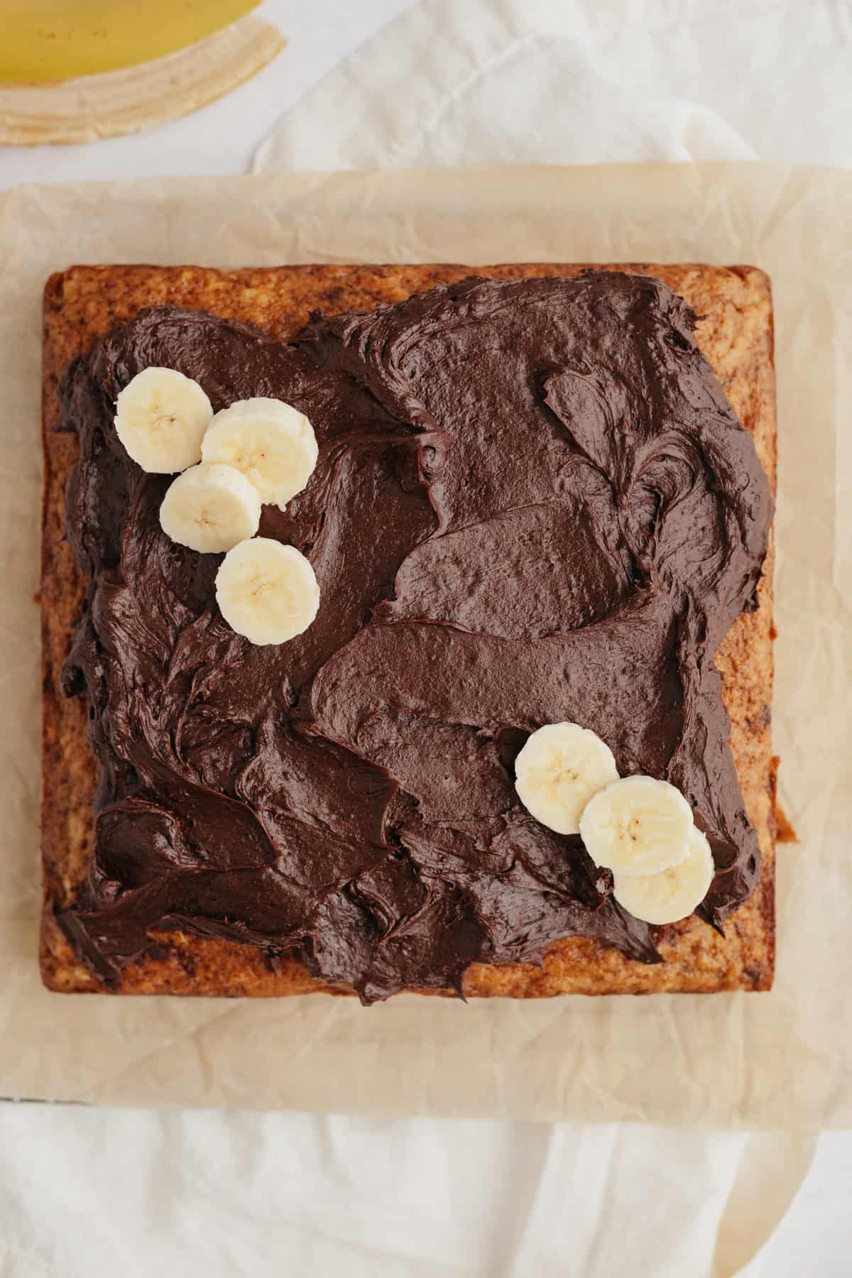 A square banana cake topped with chocolate frosting and banana slices.