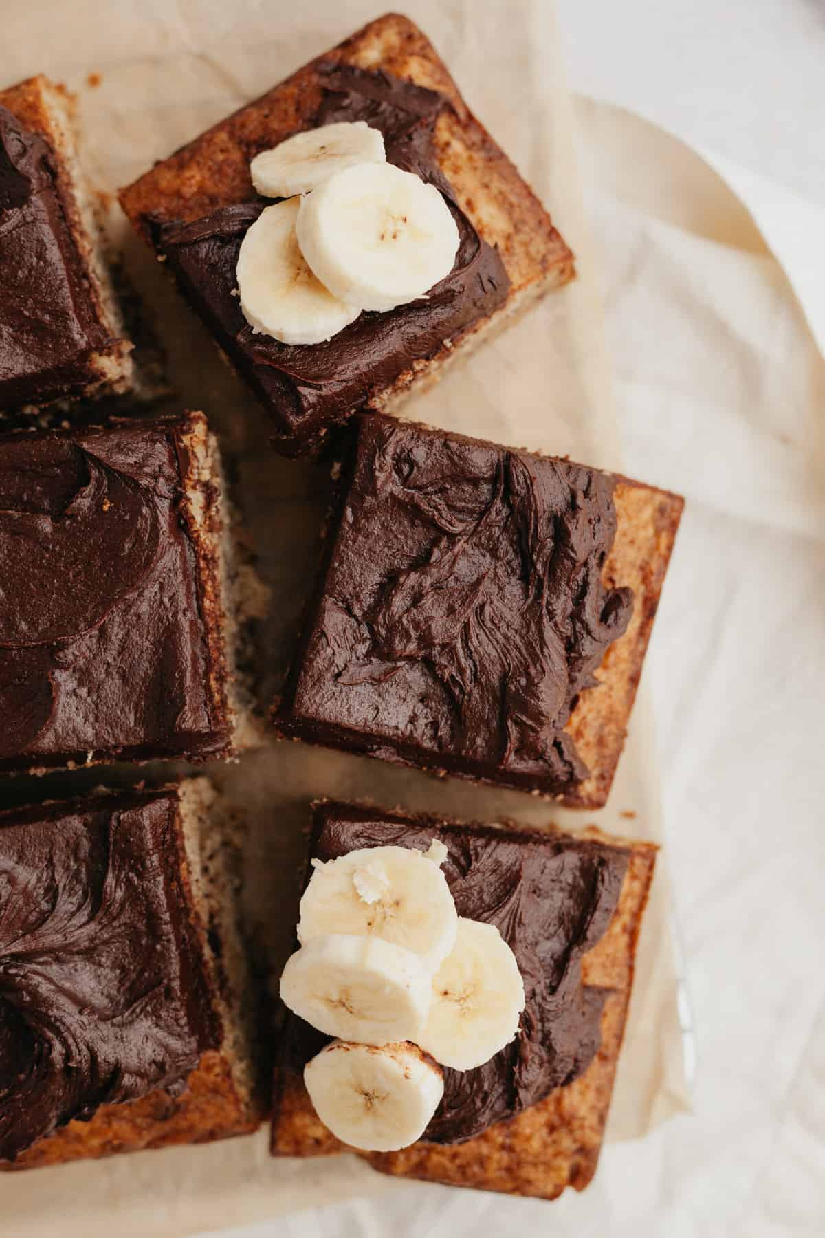 Squares of banana cake with chocolate frosting on parchment paper.