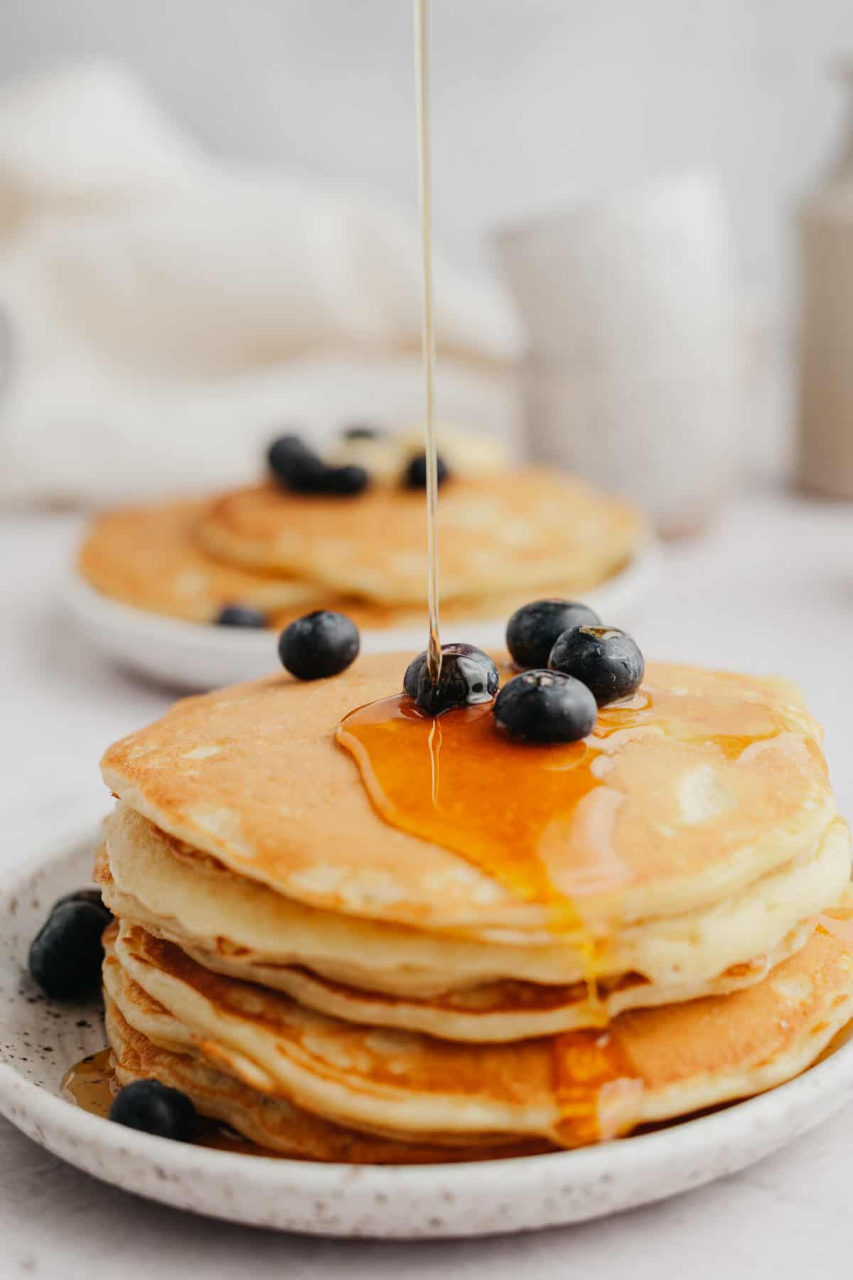 A stack of pancakes topped with blueberries, syrup is being poured on top.