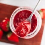 A mason jar with strawberry filling for cake with a silver spoon.