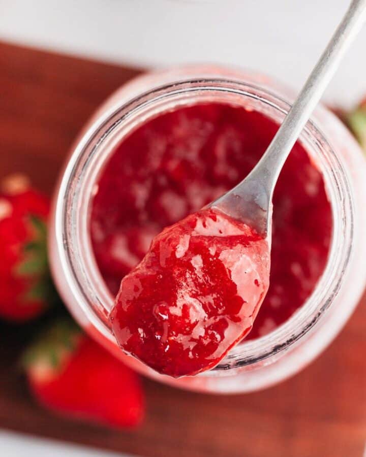 A mason jar with strawberry filling for cake with a silver spoon.