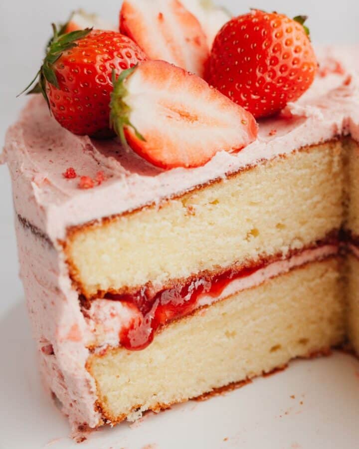 A close up of a two layer vanilla cake with a strawberry frosting, the cake has been cut.
