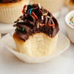 A vanilla cupcake topped with chocolate frosting and rainbow cupcakes, a bite has been taken out of the cupcake.