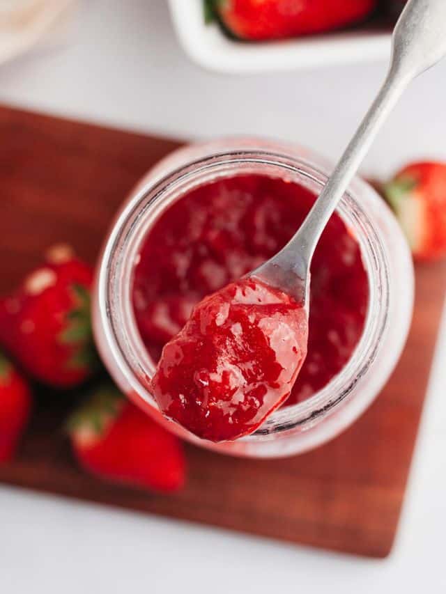 A mason jar with strawberry filling for cake with a silver spoon.