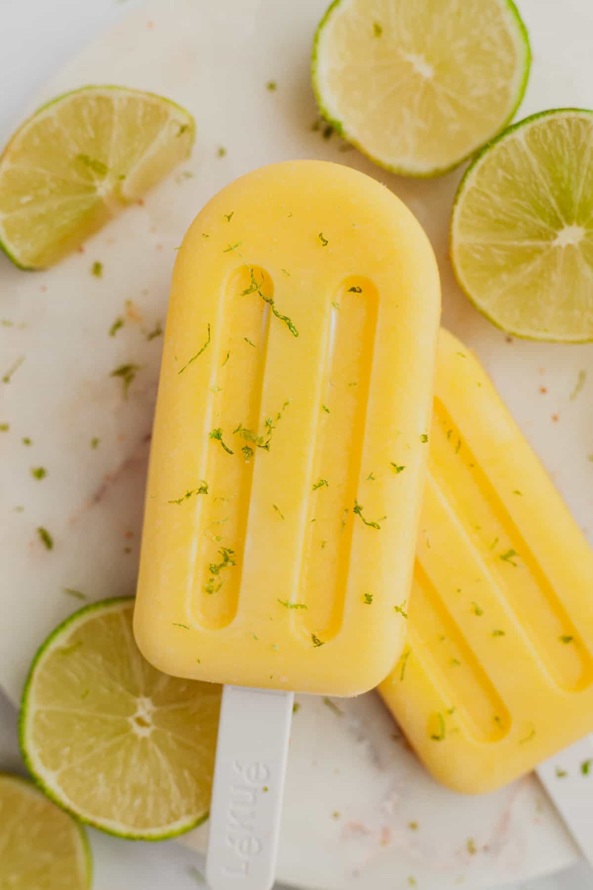 Two mango popsicles on a white plate with some slices of lime.