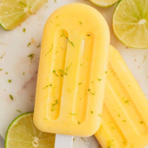 Two mango popsicles on a white plate with some slices of lime.