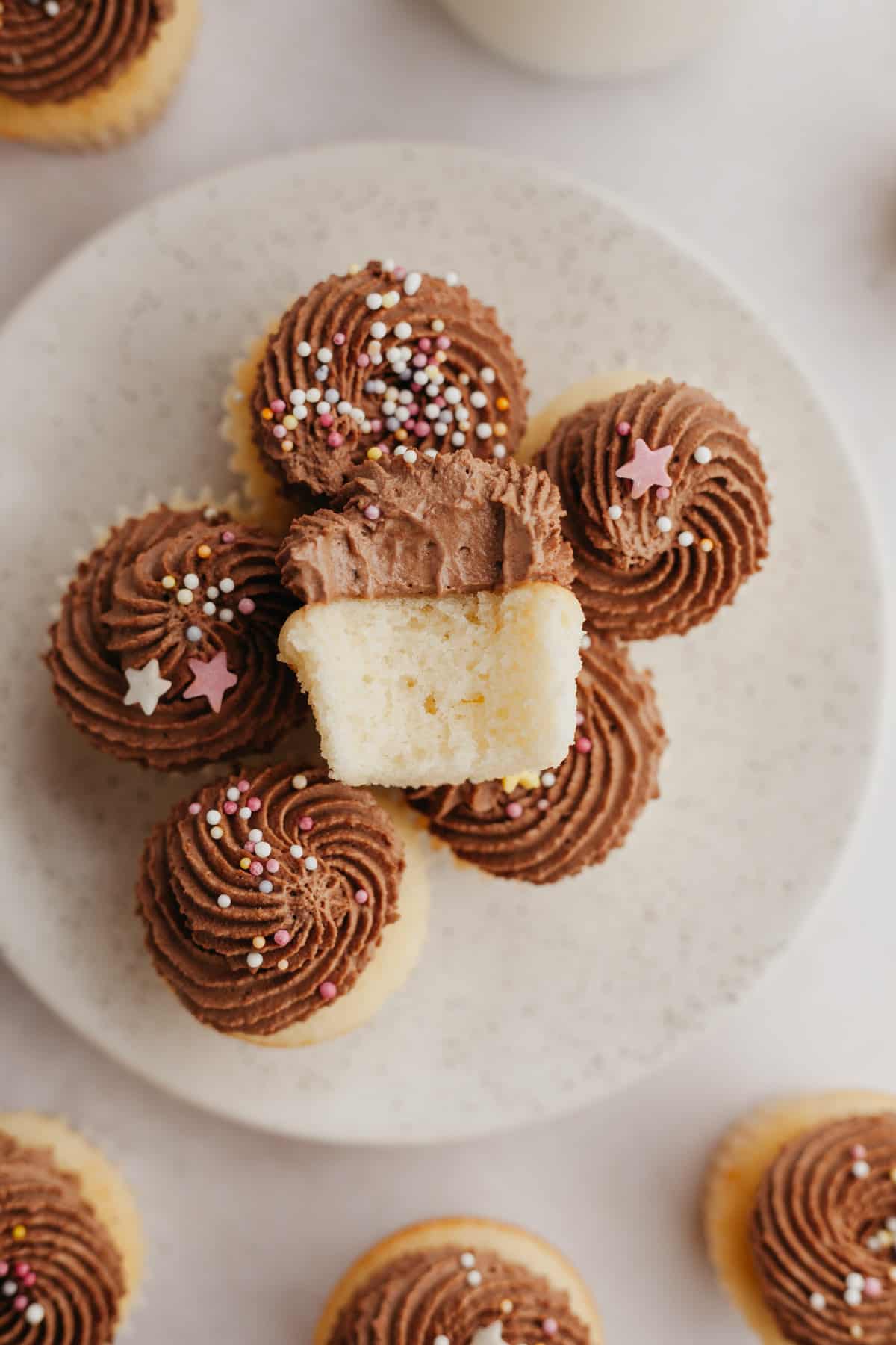 A bunch of mini vanilla cupcakes with chocolate frosting on a plate, one cupcake is on top with a bite taken out of it.