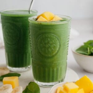 Two green smoothies in glasses, one has mango pieces on top.