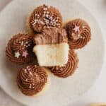 A bunch of mini vanilla cupcakes with chocolate frosting on a plate, one cupcake is on top with a bite taken out of it.