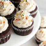 Chocolate cupcakes with a white frosting and rainbow sprinkles on a cake stand.