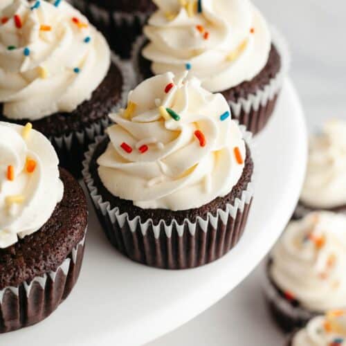 Chocolate cupcakes with a white frosting and rainbow sprinkles on a cake stand.